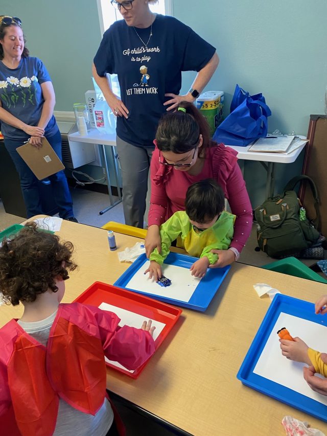 Parents help their toddlers paint while ECI Playgroup Instructors watch.