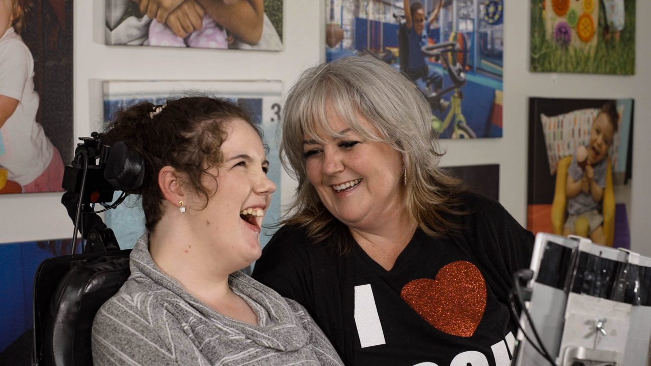 Megan, who uses a wheelchair with AAC tablet attached, and her mom.